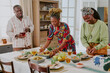 © AnnaStills - Three Black adults preparing traditional Kwanzaa meal at dining table, woman arranging food on platter, man pouring drink, another woman placing fruit, festive family celebration scene