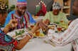 © AnnaStills - Group of middle aged Black adults sitting at table holding hands and praying during Kwanzaa celebration, traditional kinara with candles and festive food visible in background
