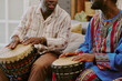 © AnnaStills - Two Black men playing traditional drums during Kwanzaa celebration, sitting side by side, focusing on rhythmic hand movements, wearing patterned clothing, engaging in cultural activity
