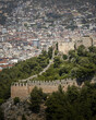 View of ancient stone walls snaking along a verdant hillside, contrasting with the dense cityscape stretching towards the distant horizon, Alanya, Antalya, TÃ¼rkiye.