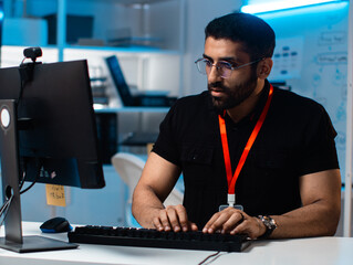 Focused Male IT Professional and Software Programmer Writing code using desktop computer review and analysis data on monitor screen developing a new software app development project in office night.