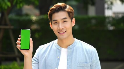 Cheerful young Asian man holding a smartphone with a blank green screen, looking at the camera for a technology advertisement