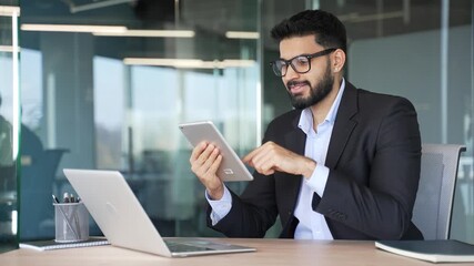 Smiling businessman is using browsing digital tablet sitting at workplace in business office. Worker in formal suit is chatting online, working in app, reading message or chatting online with client
