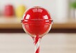© anzuragfx - Closeup of a bright red, round lollipop with a white and red striped stick, against a blurred background of colorful objects