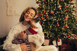 © polinaloves - Young beautiful woman and her cute dog sitting under Christmas tree in a decorated living room.