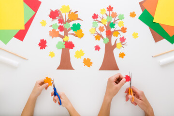 Wall Mural - Mother and child hands cutting maple leaves on light gray table background. Creating trees with colorful leaves from application paper. Closeup. Making autumn decoration. Flat lay. Top down view.