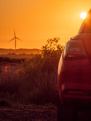 Wall Mural - A red car is parked in a field with a sunset in the background. The car is facing the sun, and the sky is orange and pink. Wind farm turbines in the background. Sightseeing modern technology concept