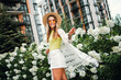 Young woman in sunhat smiles strolls through sunny park white hydrangeas beside modern city buildings and a stylish white fringe cardigan