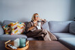 © okrasiuk - Middle aged smiling woman sitting on sofa and looking away while drinking coffee or tea. Mature senior woman relaxing after housekeeping, home cleaning. Portrait of relaxed female resting at home.