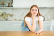 © JackF - In kitchen with light furniture and daylight, long-haired girl in white T-shirt put elbows on table and props chin with fist. Home comfort concept