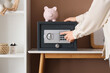 © Pixel-Shot - Woman with piggy bank opening modern safe box on table near brown wall, closeup