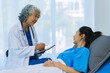 © ArLawKa - Elderly female doctor and Asian female patient are discussing something while sitting on hospital bed. Medical consultation concept.