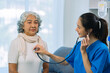 © ArLawKa - Senior Asian female patient listening to doctor using stethoscope in hospital. Doctor in uniform using stethoscope to examine heart. Senior woman.