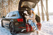 © maxbelchenko - A cheerful woman sits in trunk of a car in snowy forest with her pet. A woman and her Siberian husky are on a road trip on a sunny winter day. Concepts include adventure, relaxation, and weekends.