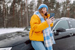 © maxbelchenko - Portrait of happy woman in a bright jacket taking selfie while standing next to car on winter day. Beautiful woman with phone in a snowy forest, enjoying nature. Concept of transportation, travel.