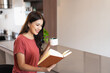 © Fahng - Young woman drinking tea and reading book in her kitchen while enjoying morning at home.