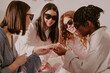 © AnnaStills - Group of diverse young adult women wearing sunglasses celebrating bachelorette party, smiling and laughing while looking at each others hands in bright indoor setting