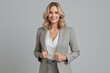© The 2R Artificiality - Portrait of a woman with blonde hair wearing a light gray business suit standing against a gray background smiling confidently with elegant attire and casual style.