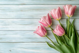 A cluster of  soft pink tulips with bright green leaves lies diagonally across a background of weathered, light color wooden planks, providing copy space.