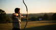 © haris - A man draws back a bow aiming at a target on a field, backlit by the sun.