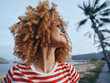 © SHOTPRIME STUDIO - Closeup portrait of a joyful person with thick curly hair, sunglasses, and a striped shirt, looking upward with a bright mood against a beachside backdrop.