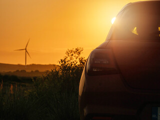 Wall Mural - A red car is parked in a field with a sunset in the background. The car is facing the sun, and the sky is orange and pink. Wind farm turbines in the background. Sightseeing modern technology concept