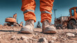 © Oleksandr - Construction worker in personal protective equipment stands on a job site with heavy machinery. Work boots on gravel, orange work pants visible.