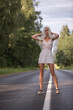 © shymar27 - Female model with long blonde hair wearing a white dress stands confidently on a deserted road surrounded by lush greenery, embodying freedom and adventure in nature