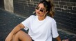 © YOUSSEF KETTOU - Confident young woman with long hair in a ponytail wearing sunglasses and a plain white oversized t-shirt, posing against an urban dark brick wall. Street style fashion apparel mockup with copy space