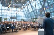 © andyaziz6 - A speaker addresses a large audience in a modern building with natural light and high ceilings.