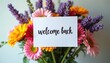 © Pete - Colorful flower bouquet. White card with handwritten phrase welcome back. Gerbera daisies roses are in full bloom. Greeting concept for a message.
