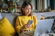 © VolumeThings - Young Asian woman online shopping with laptop smartphone and credit card in living room