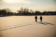 © StockMediaSeller - Two people walking across a snow-covered frozen lake at sunset. Their long shadows stretch across the sparkling snow, creating a peaceful winter landscape with a golden evening light.