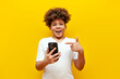 © Богдан Маліцький - shocked african american boy using smartphone and pointing at copy space on yellow isolated background, surprised schoolboy teenager holding mobile phone and pointing at empty space in amazement