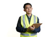 © yashis007 - Confident Asian male engineer or construction site manager in a safety vest holding a clipboard, looking up thoughtfully against an isolated white background