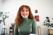 © Studio Marmellata - A young woman with curly red hair smiles warmly while looking at the camera. She is wearing a green turtleneck sweater and holding a notebook.