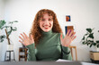 © Studio Marmellata - A young woman with curly red hair smiles and waves hello while working from home.