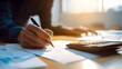 © rehan - Person's hand writing on paperwork with a pen, alongside a calculator on a wooden surface in a brightly lit setting.