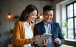 © zhao - Young cheerful asian woman showing something on digital tablet to her male colleague while standing together in the coworking space or office. High quality