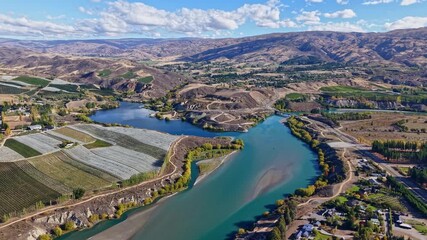 Wall Mural - Drone ascends and pulls back above turquoise river, bridge, and farmlands under blue sky.
