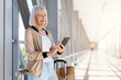 © Anastasiya - Mature woman standing at airport corridor with luggage using smartphone