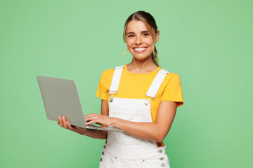 Young happy IT woman she wear yellow t-shirt overall casual clothes hold use work on laptop pc computer chat online isolated on plain pastel light green background studio portrait. Lifestyle concept.