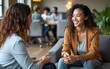 © fu - Smiling young multiethnic woman talking to senior female colleague exchanging work experience or assisting coworker with professional development, while sitting in lounge area of coworking space