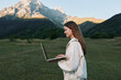 © SHOTPRIME STUDIO - A cheerful female professional works on a laptop outdoors in a scenic mountain meadow. She is smiling and focused, creating an inspiring productive outdoor workspace.