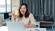 © amnaj - Asian woman smiling, enjoying a video call with a laptop, holding a mug, working from her modern home office