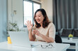 © amnaj - Asian woman smiling and waving at laptop during a remote video meeting from her home office, confident and engaged in conversation