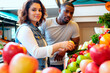 © pressmaster - Caucasian young adult woman and Black young adult man selecting fresh produce at market stall, both looking at camera, surrounded by colorful fruits and vegetables, casual setting