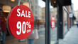 © Viktor - Red circular sign advertises 90 off sale in shop window. Another sign visible on store window reflects street and passersby. Customers rush to get deals.