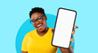 © Prostock-studio - A woman with short curly hair wears glasses and a bright yellow shirt. She smiles widely while holding a blank mobile device in front of her against a vibrant blue backdrop.