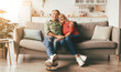 © Prostock-studio - Happy Mature Couple Embracing Posing Sitting On Couch At Home, Smiling Looking At Camera. Retirement And Family Lifestyle, Marriage And Happiness Concept